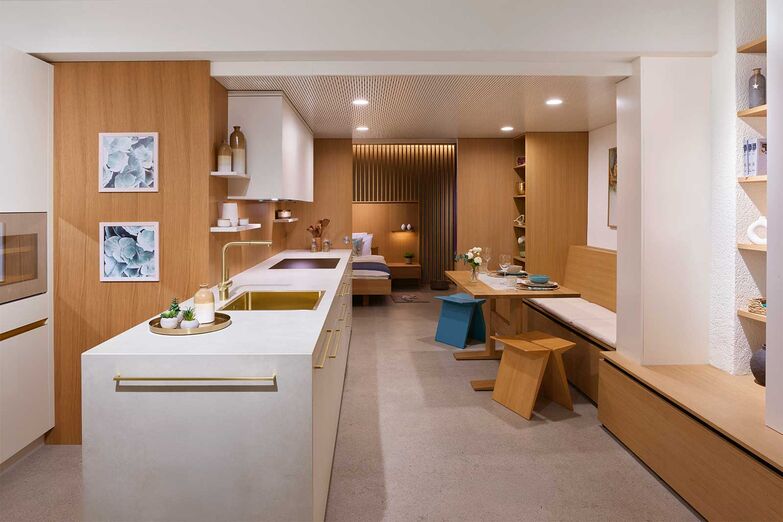 Einrichtung für ein Tiny House: Küche, Essplatz, Stauraum, Schlafzimmer und Bad finden auf kleinstem Raum Platz und bieten ein luftiges und funktionales Ambiente. Die Küche besticht durch einen hellen, warmen Farbton und harmoniert mit der fast gleichfarbigen Keramikabdeckung. Die Griffe, die Mischer und das Becken in zartem Gold sowie die Einbaugeräte und der Weinkühler in der Farbe Pearl verleihen einen Hauch von Luxus. Das Holzfurnier für Wände und Schiebetüre kann in der Produktion und Ausstellung in Wyssachen besichtigt werden. Die zweite Ausstellung befindet sich in Langenthal.   Loosli AG, www.loosli.swiss
